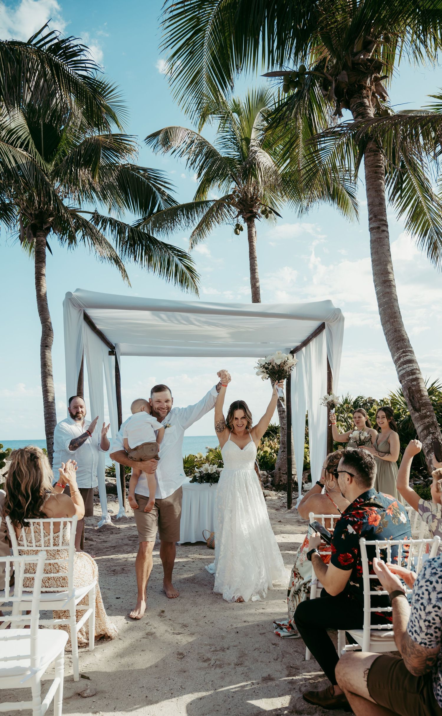 Beautiful tropical beach wedding ceremony under palm trees with white canopy and guests seated in white chairs.