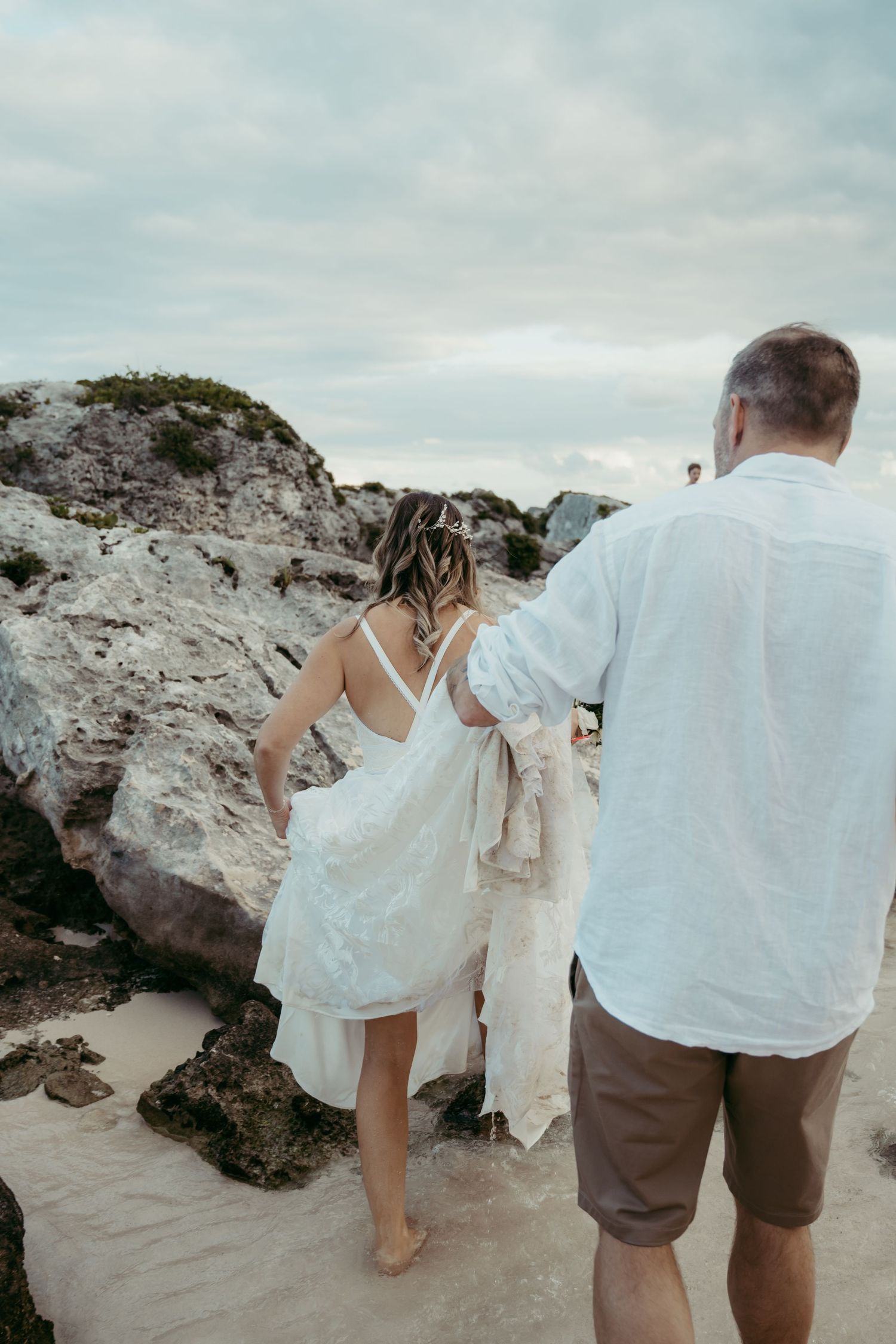 A couple in white exploring rocky coastal terrain at sunset, with the person in front walking barefoot on the beach.