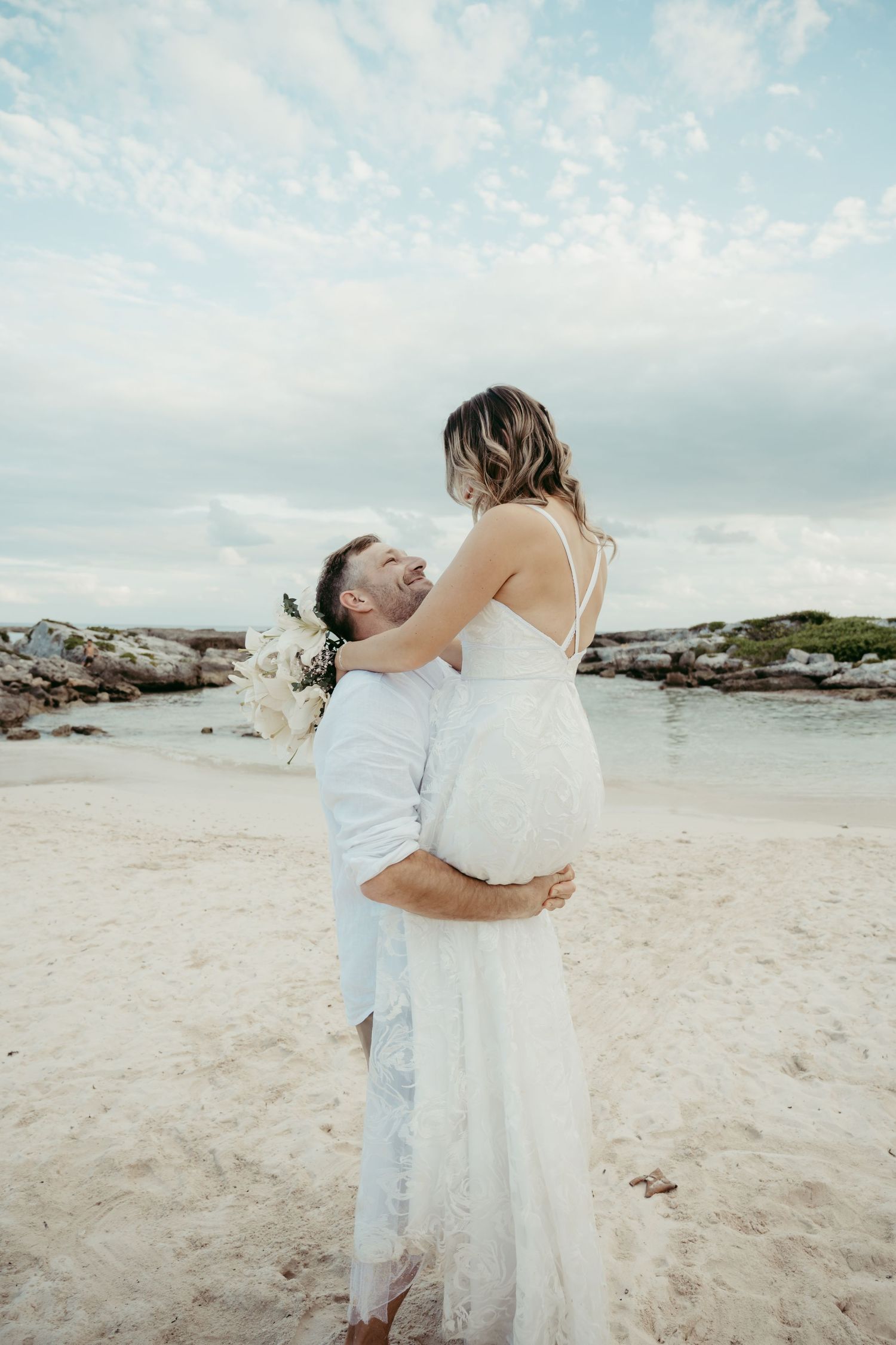 A couple embraces on a scenic beach with rocky coastline and cloudy sky during a romantic wedding photoshoot.
