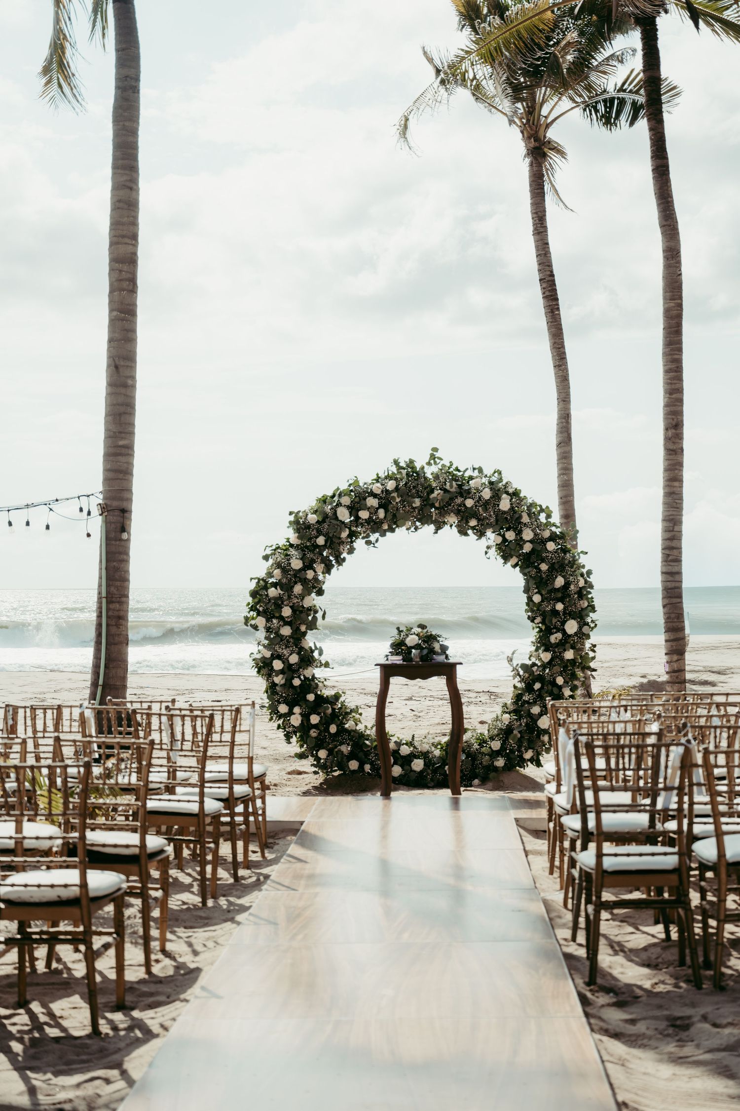 Elegant beach wedding ceremony setup with a circular floral arch, gold chairs, and ocean backdrop under palm trees.