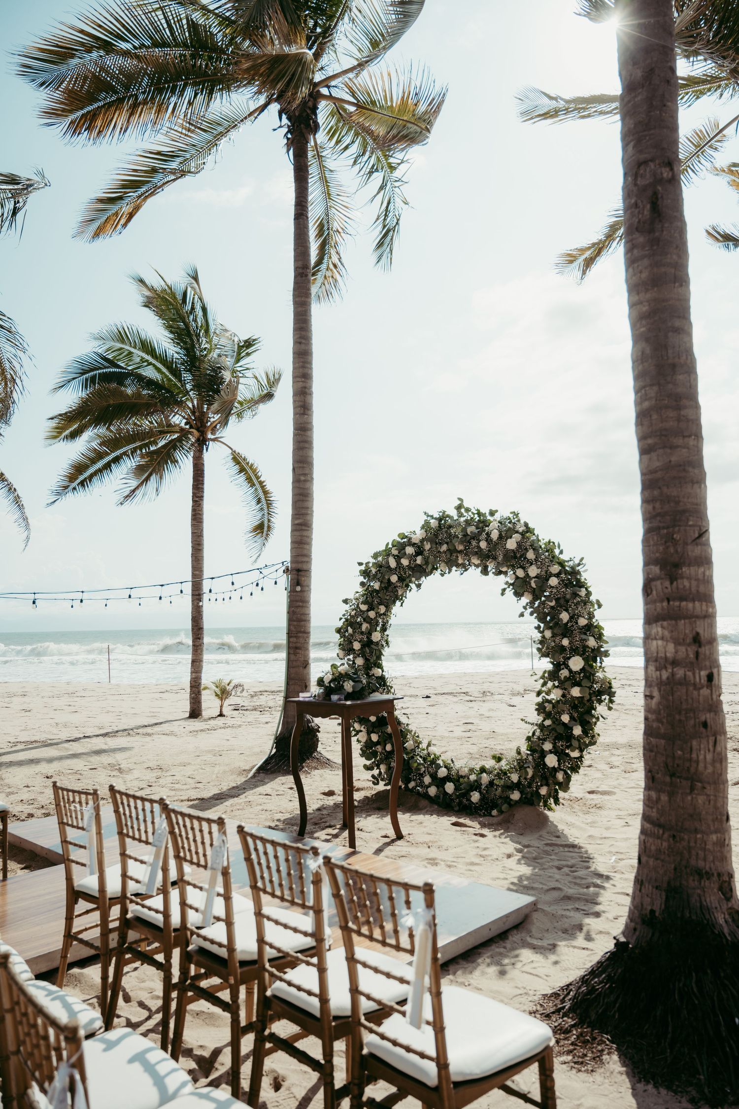 Beach wedding setup with floral hoop arch, chiavari chairs, and palm trees by the ocean.
