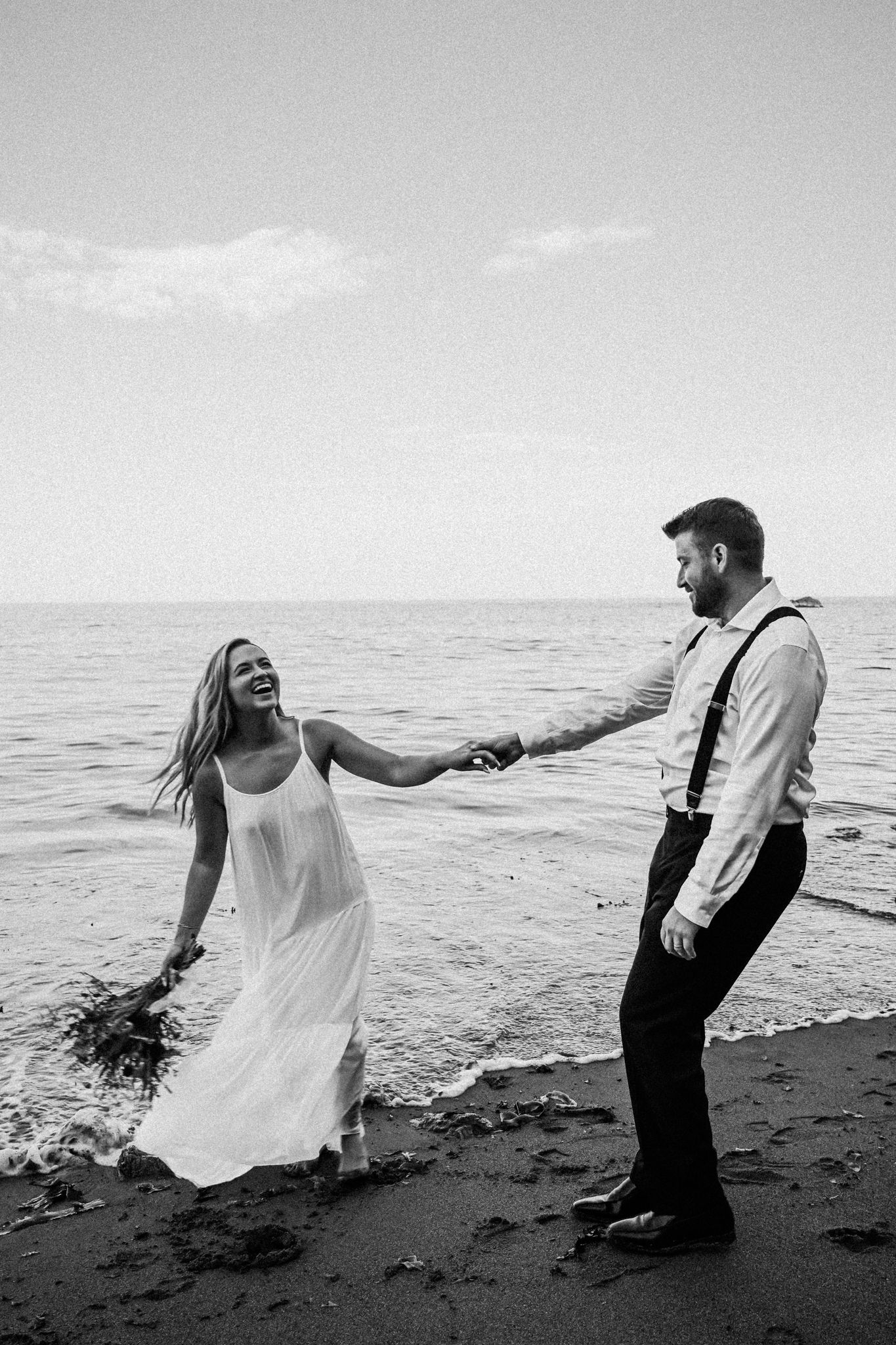 A playful couple dances on a black sand beach in black and white photography.