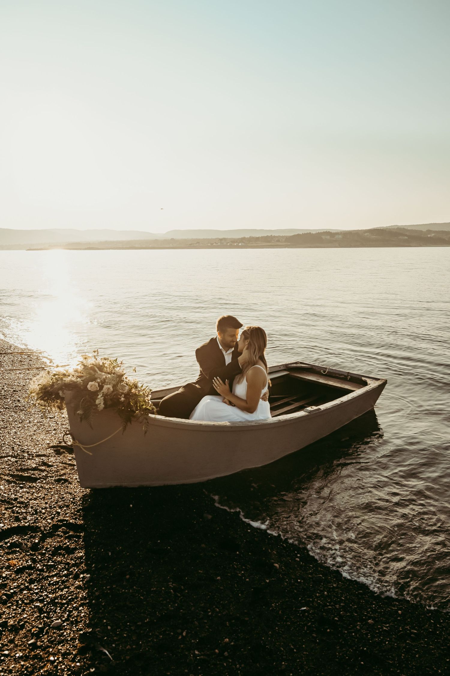 A romantic couple sits together in a wooden boat on a serene lake at sunset with flowers beside them.