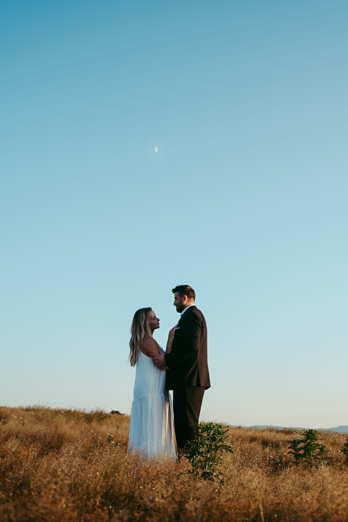 A couple in formal attire stands together in a golden field against a bright blue sky at sunset.