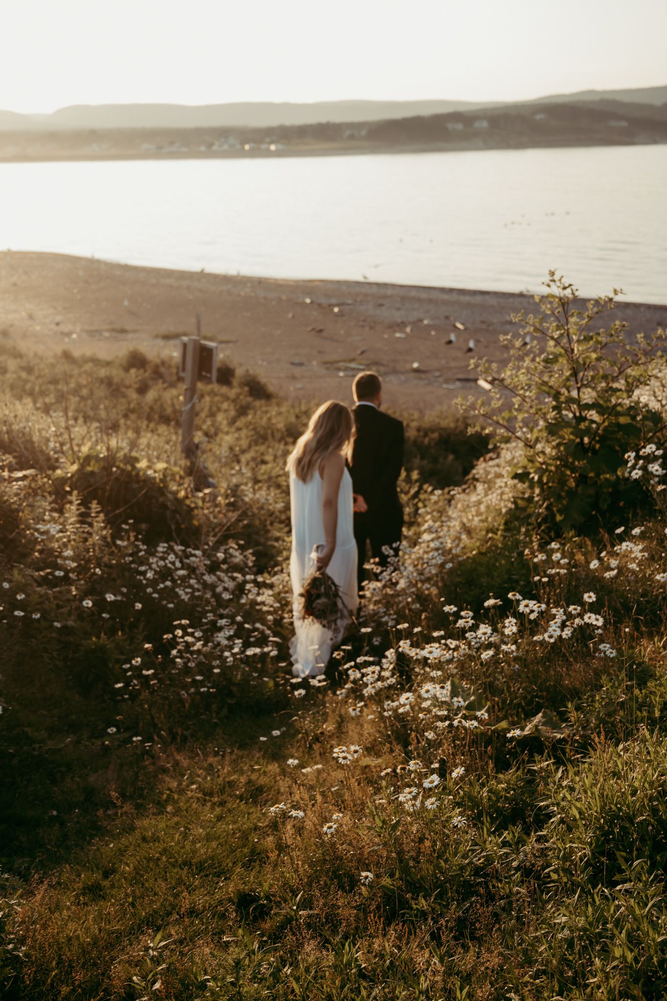 A couple walks through wildflowers on a hillside overlooking a serene lake at sunset.