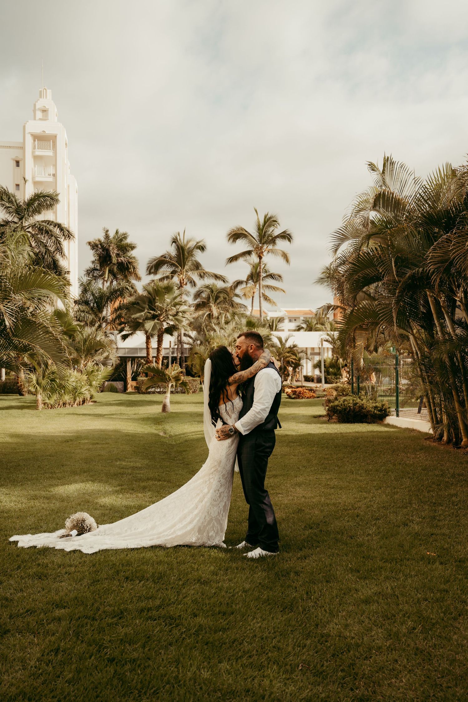 Bride and groom share a romantic kiss on lush green lawn surrounded by tropical palm trees at a resort wedding.