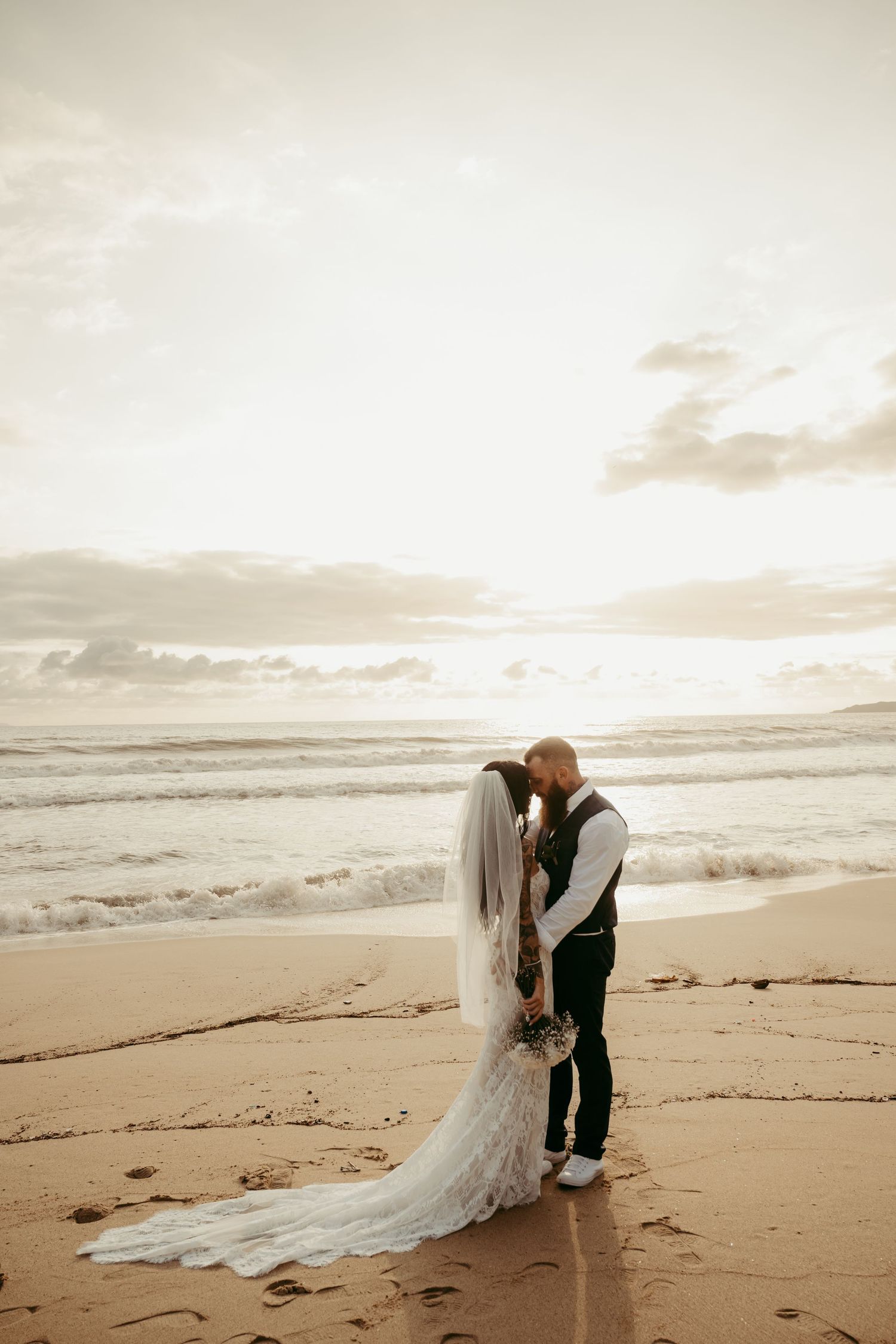 A bride and groom share a romantic moment on a sandy beach at sunset, with ocean waves in the background.