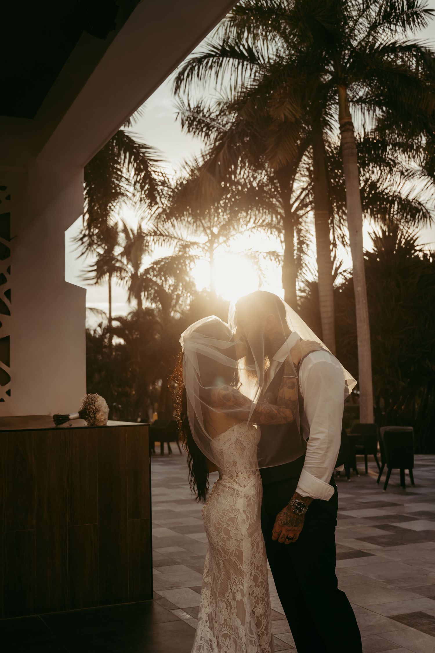 Bride and groom share a romantic sunset kiss under palm trees, the golden light glowing through her flowing veil.
