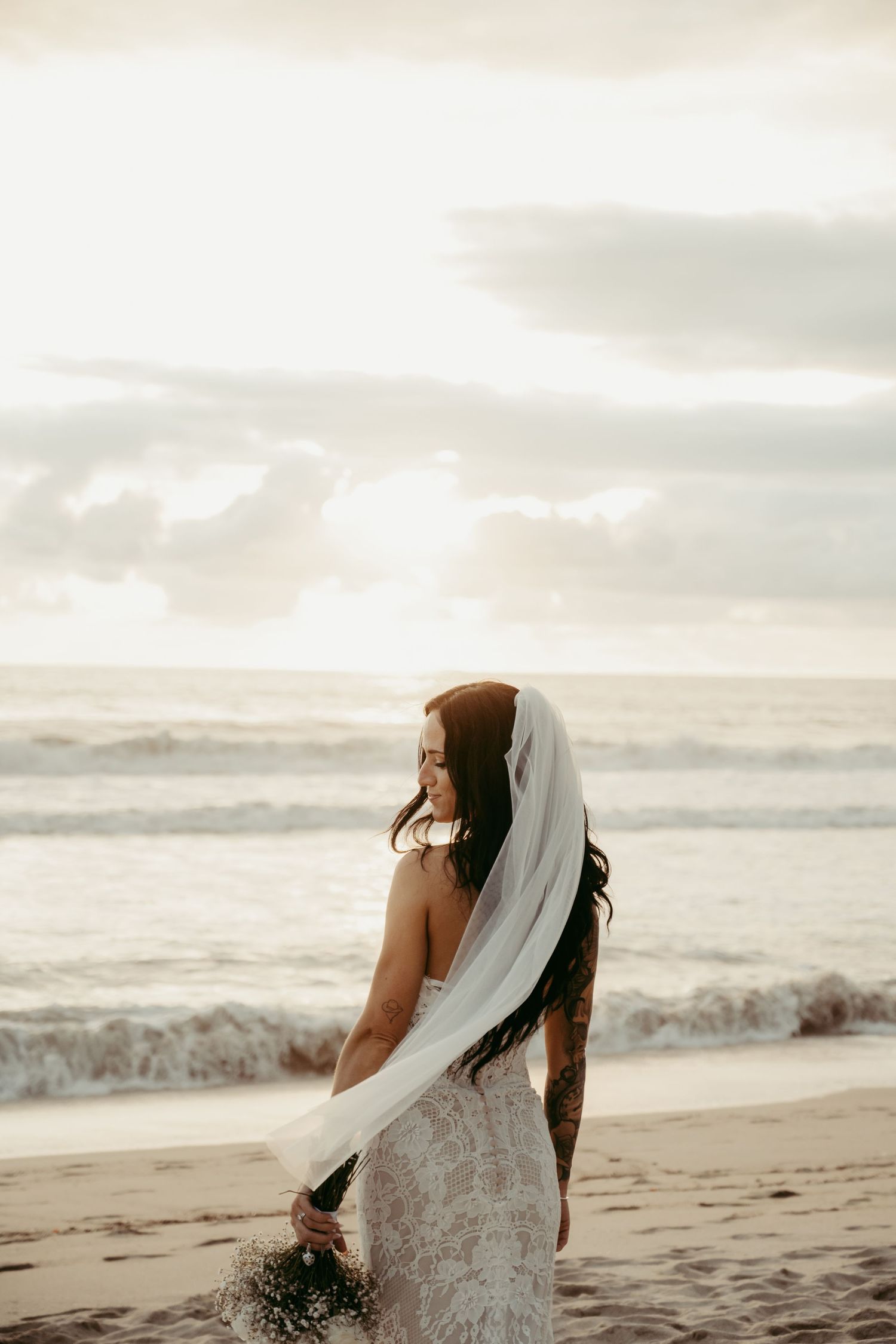 Bride in lace wedding dress and flowing veil stands on beach at sunset, tattoos visible, holding bouquet.