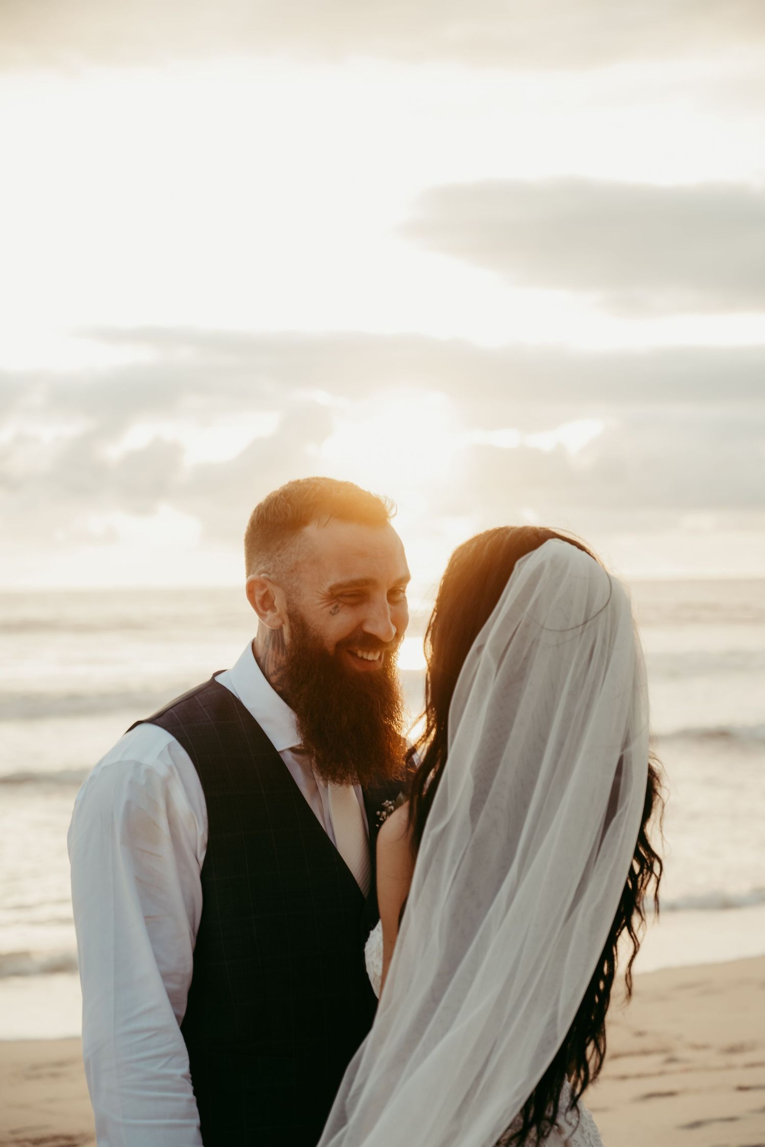 Bearded groom smiles lovingly at bride in flowing veil during golden sunset beach wedding ceremony.