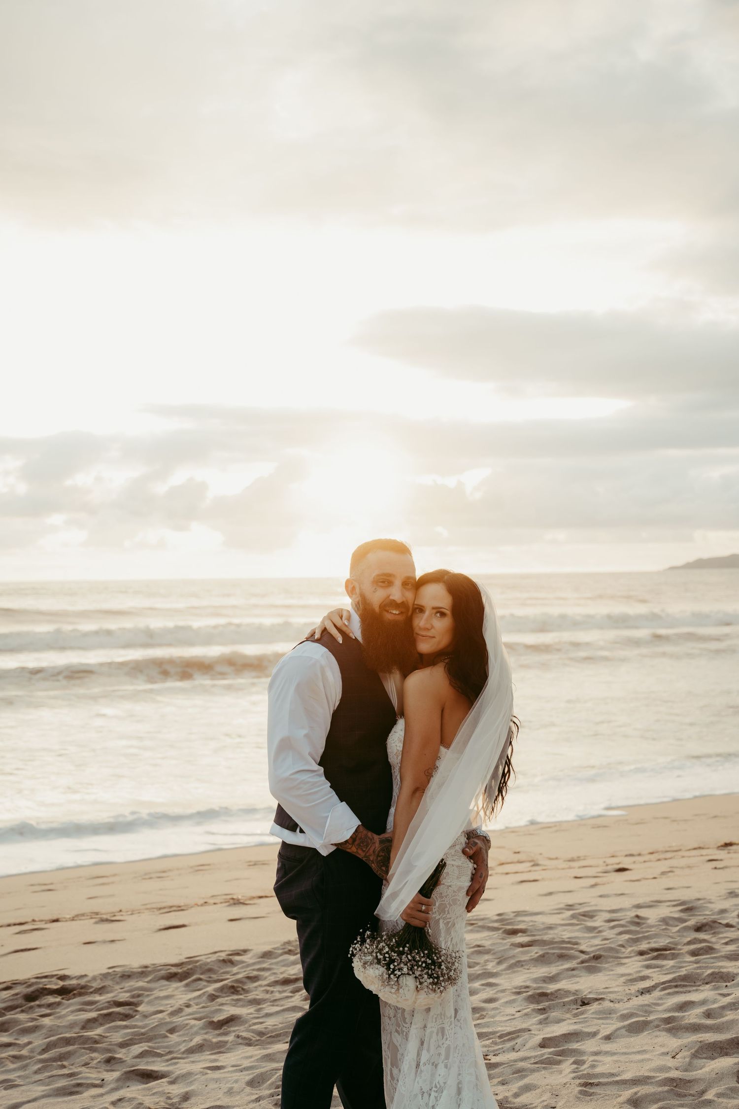Bride and groom embrace on sandy beach at sunset, bride wearing white gown with veil holding bouquet.