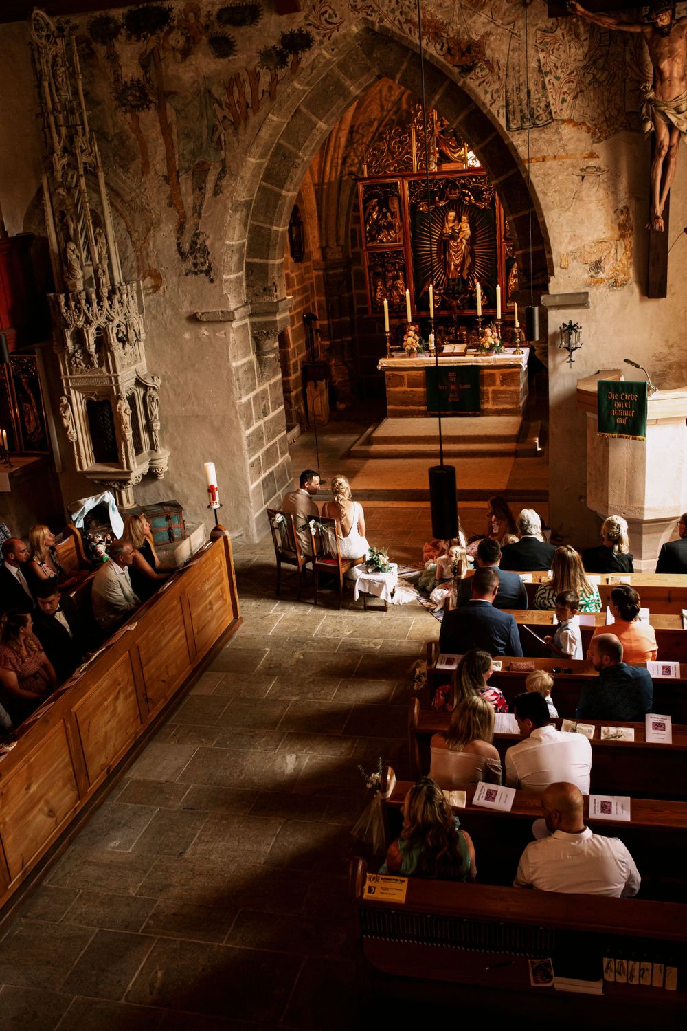 Interior of stone church during wedding ceremony with guests seated in wooden pews.