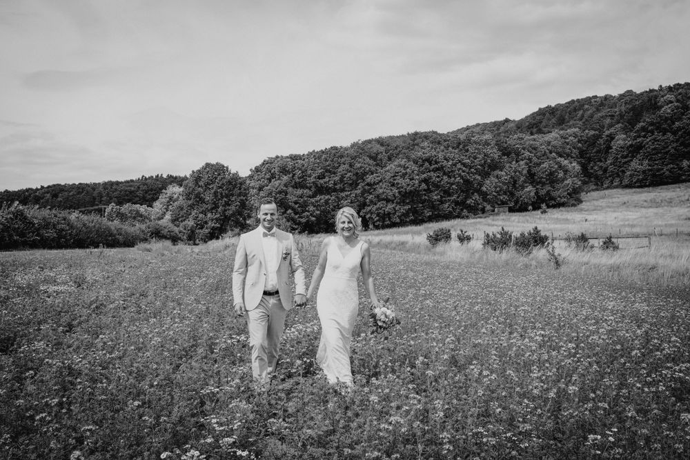 Wedding couple walking through a sunny meadow with rolling hills in the background.