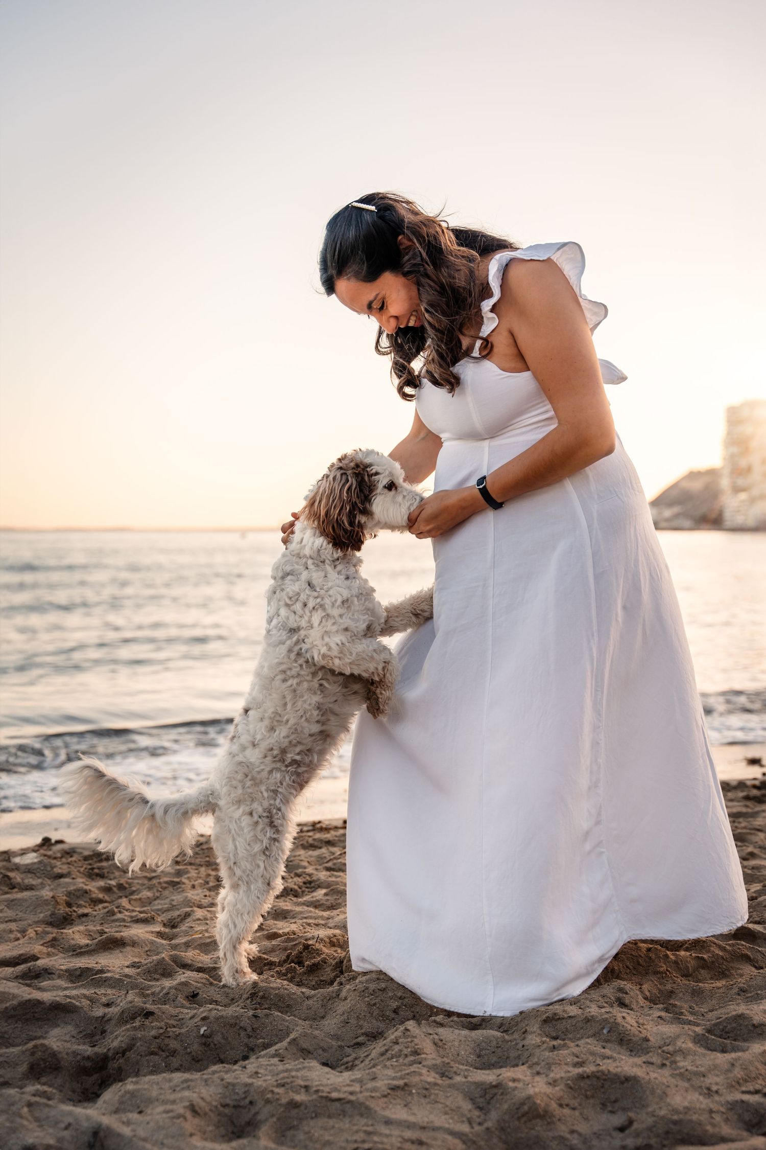 pregnant woman wearing white dress and her dog sniffing her pregnant belly while on the beach at sunset in Alicante