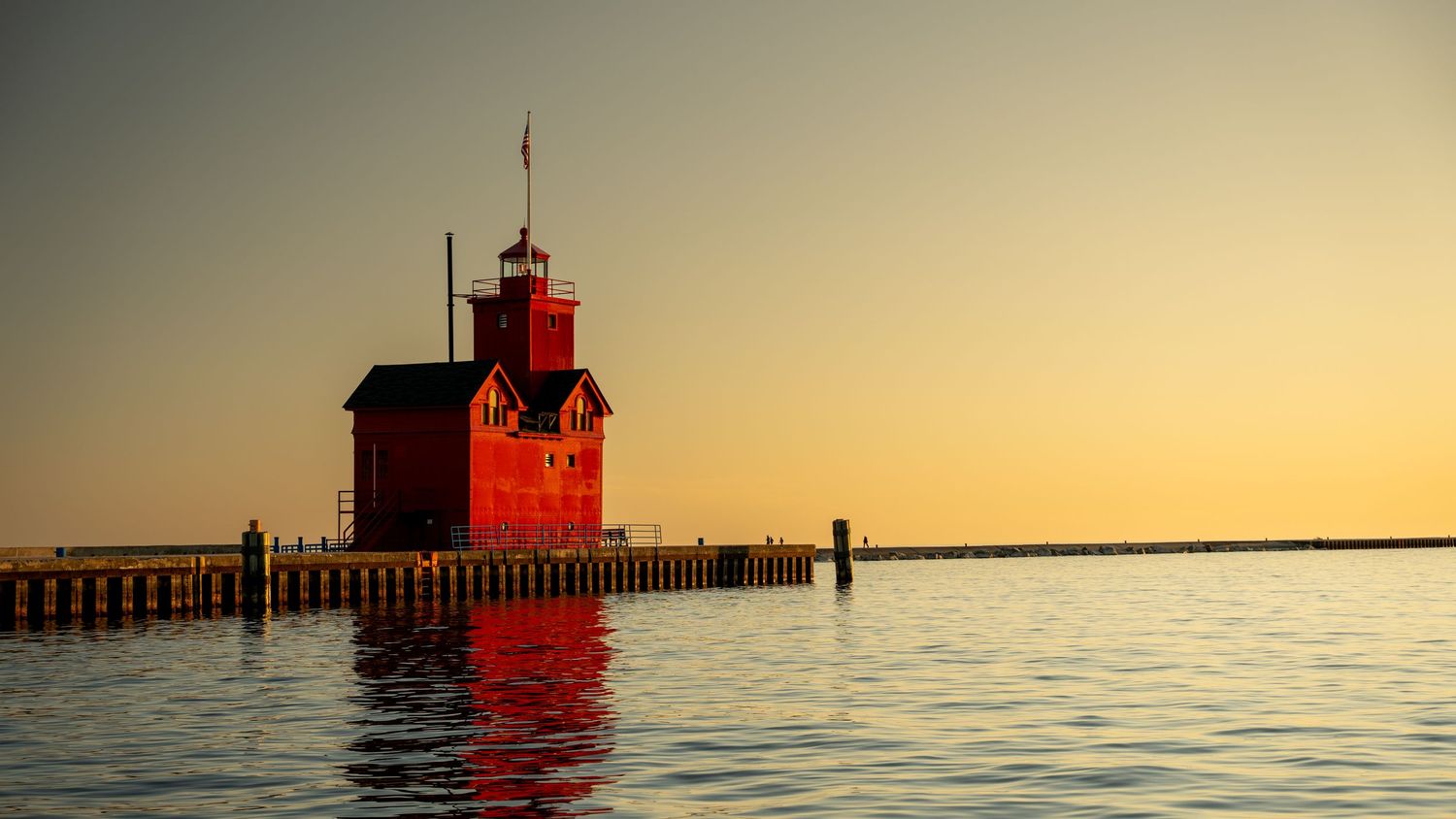 Iconic red lighthouse glowing at sunset with calm water reflections and wooden pier nearby.