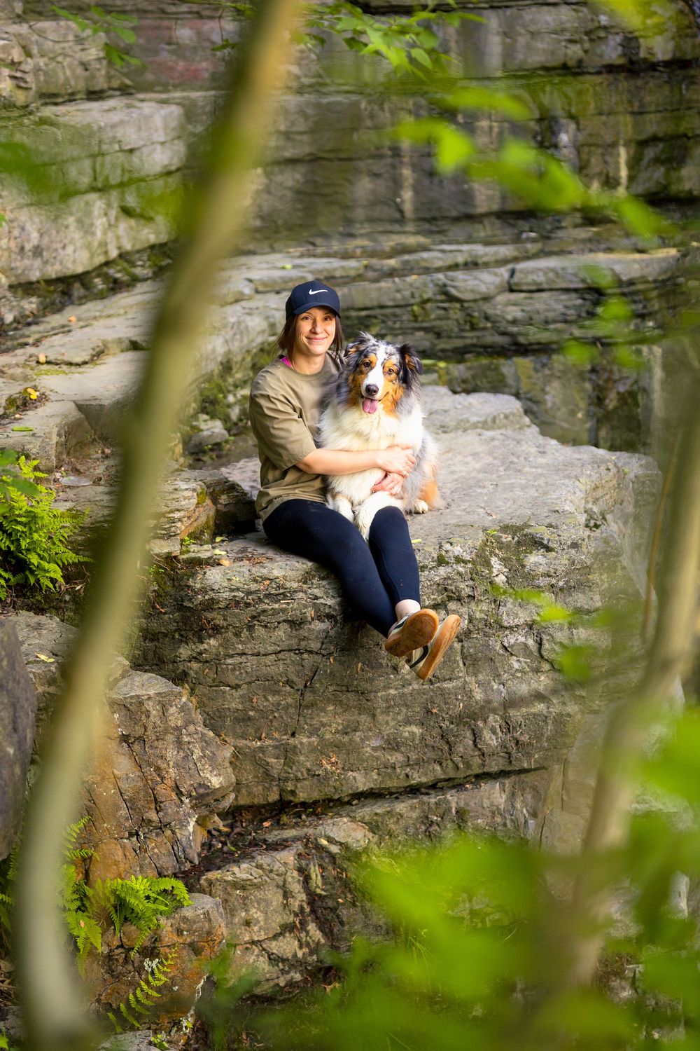 An Australian Shepherd dog and companion sit together on rocky ledge framed by green foliage in natural setting. Ottawa Pet Photographer