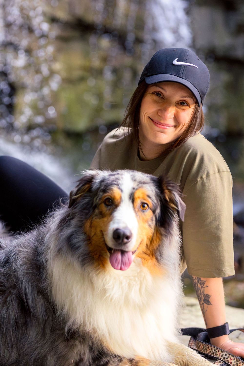 A beautiful Australian Shepherd dog with fluffy fur poses outdoors with someone wearing a cap and beige shirt. Ottawa Pet Photographer