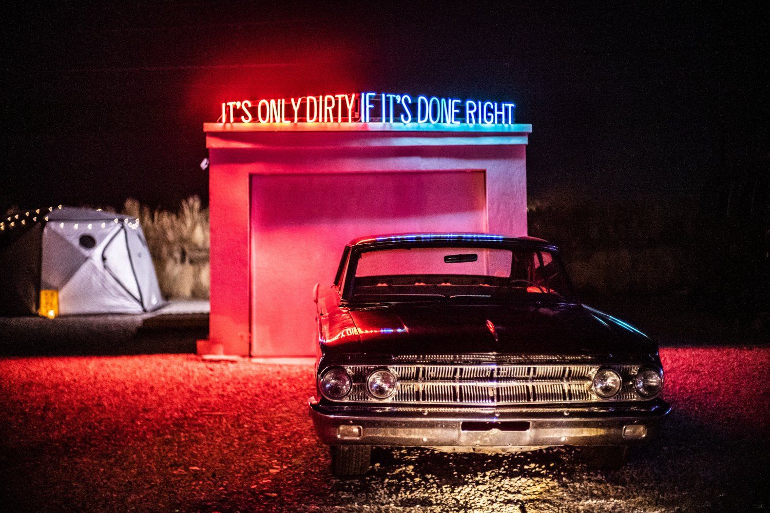 Vintage car parked at neon-lit drive-in theater entrance at night with red illumination.