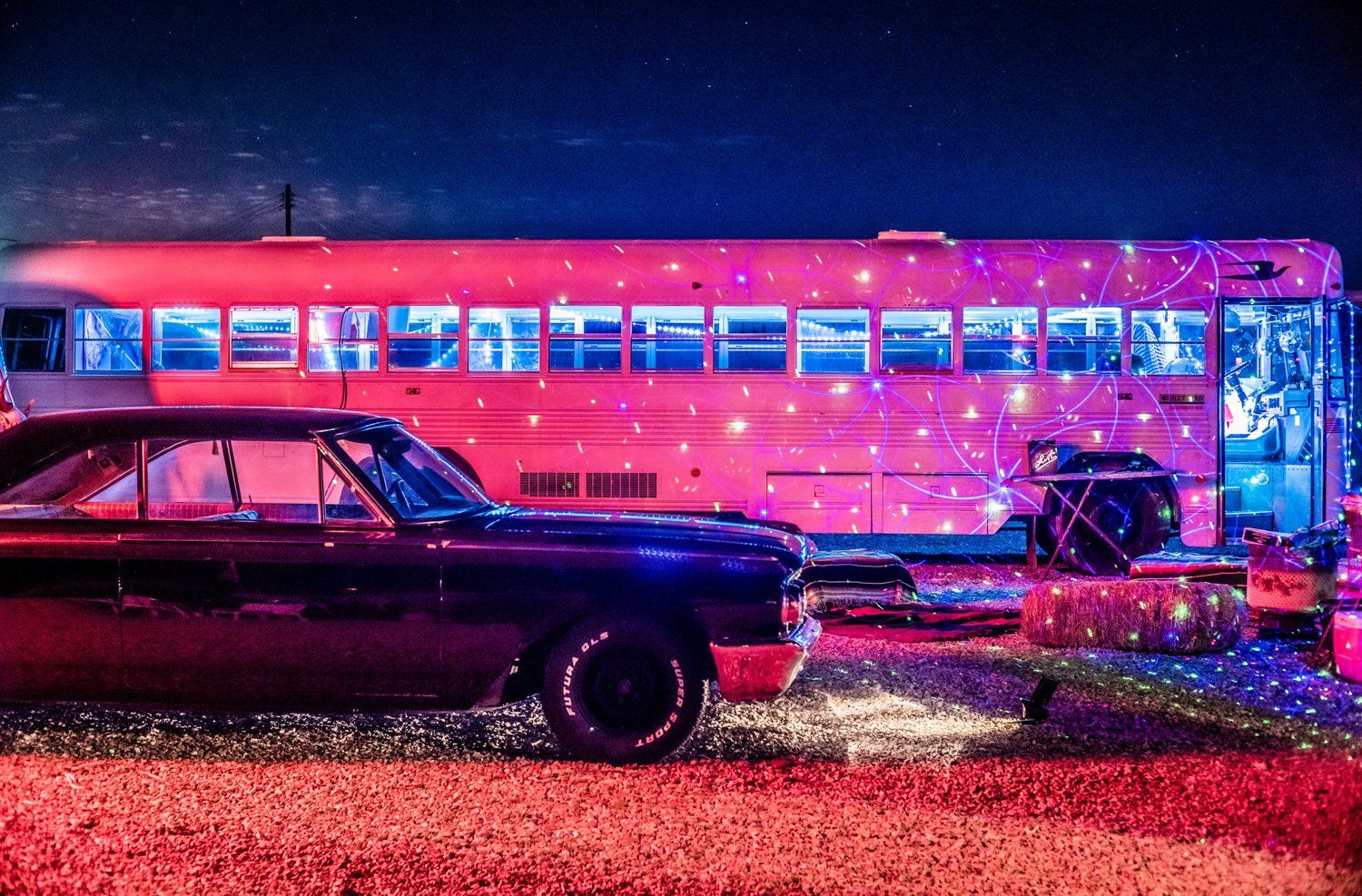 A vintage car parked next to an illuminated pink retro bus at night.