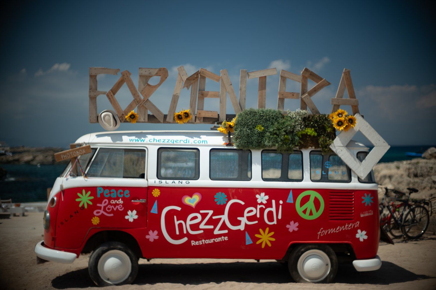 A vintage red VW bus decorated with graffiti and peace signs parked near a beach sign. Formentera