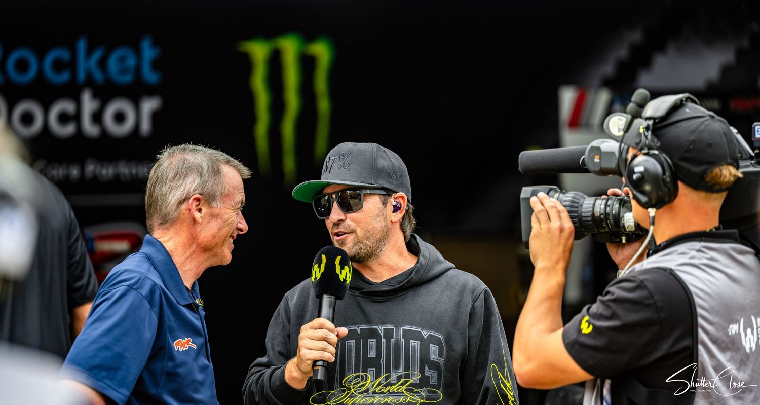 Motocross rider interviewed on camera at Monster Energy supercross event, wearing black cap and hoodie.