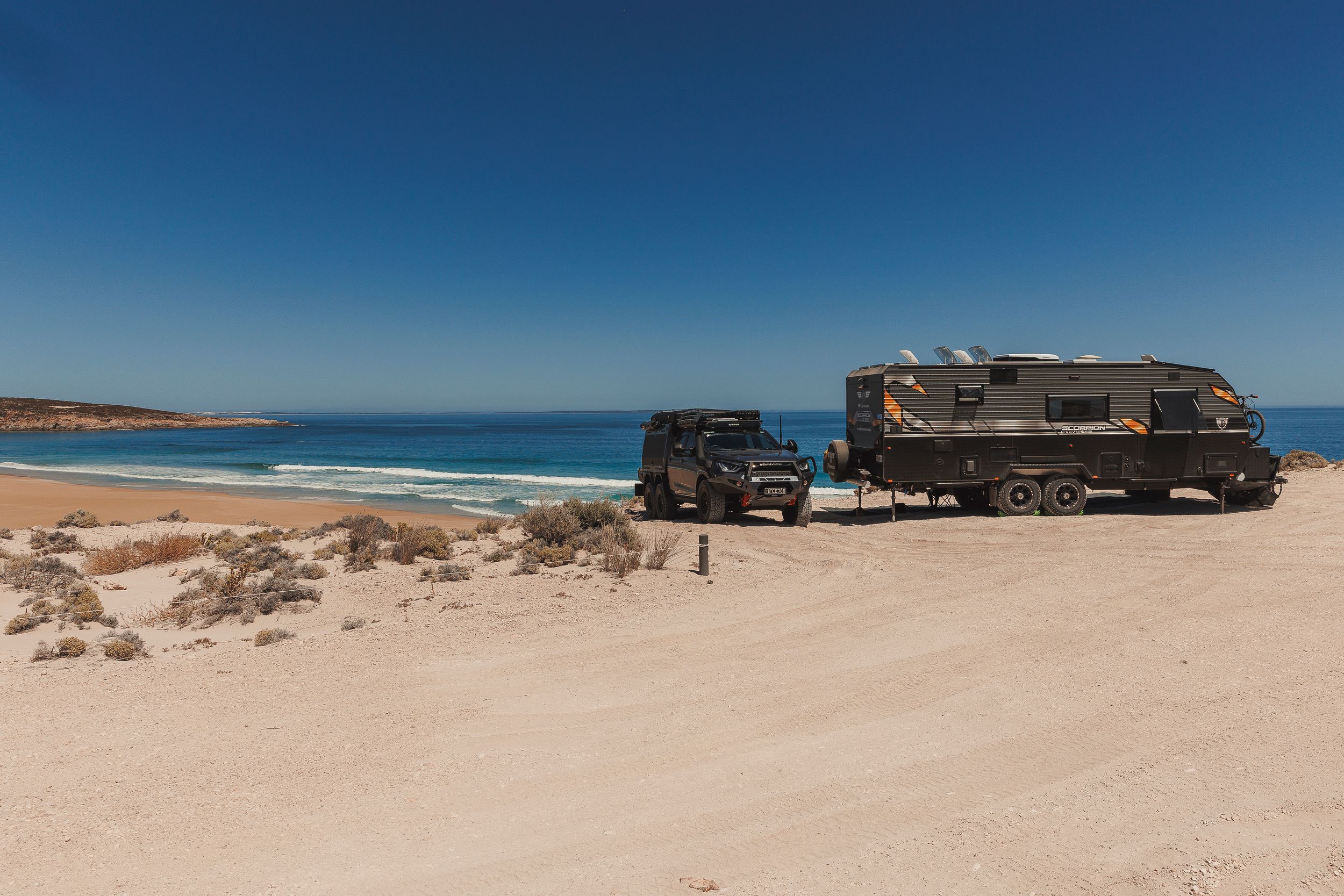 4WD vehicle and caravan parked on a sandy beach beside calm blue ocean under clear skies.
