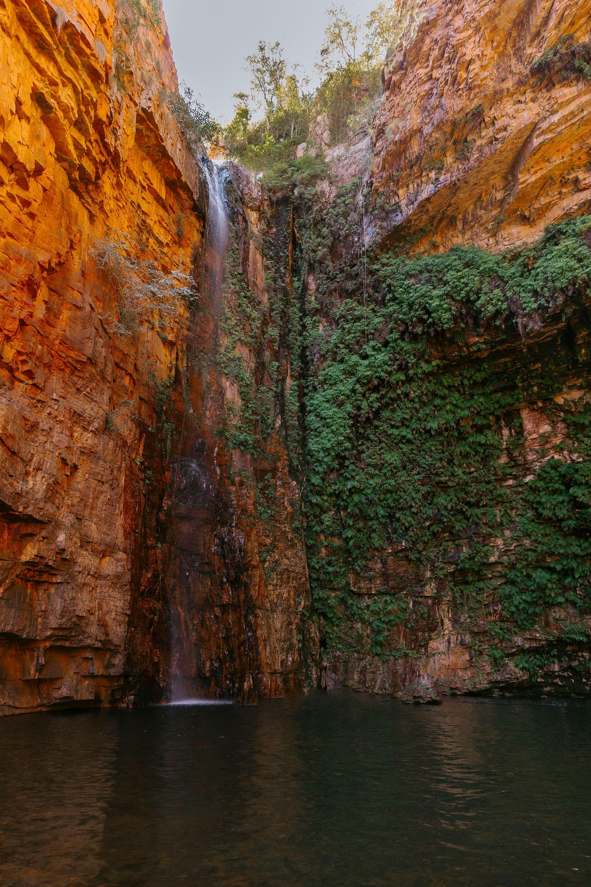 A stunning waterfall cascades down red sandstone cliffs into a dark pool, flanked by lush green vegetation in a dramatic gorge.