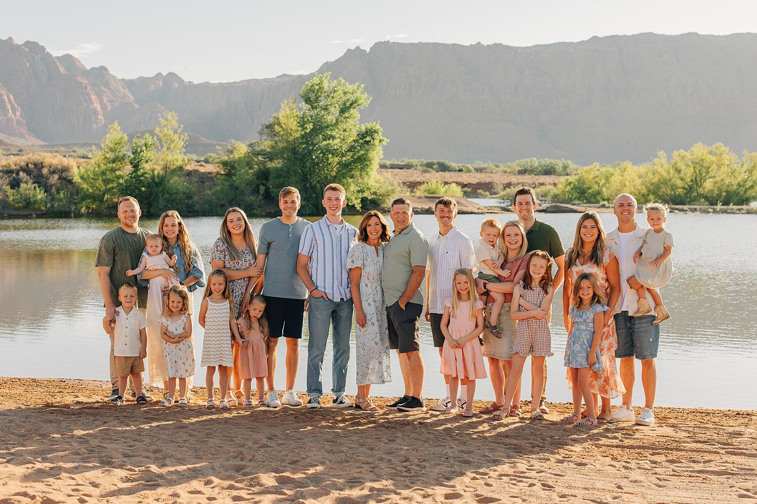 Large family group photo on sandy beach with mountain backdrop and lake during golden hour.