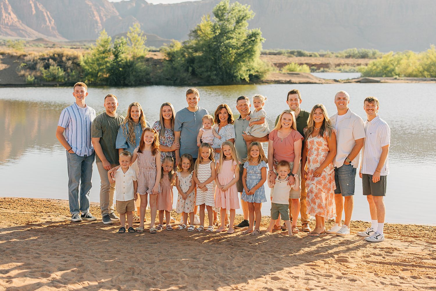 Large family group portrait outdoors near a scenic lake with red rock mountains at golden hour.