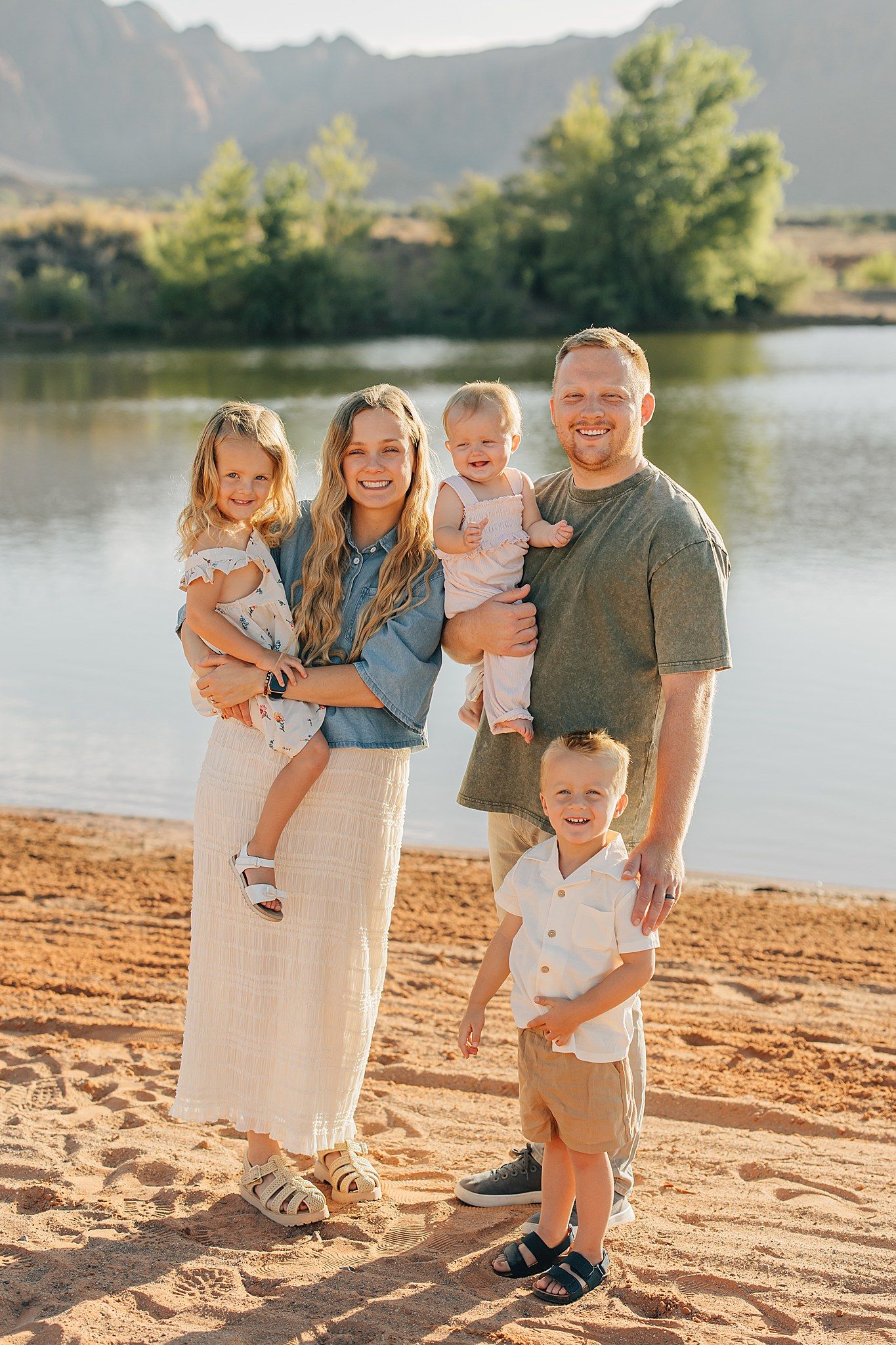 Happy family of five posing on a sandy riverbank with scenic mountain and lake backdrop in golden hour light.