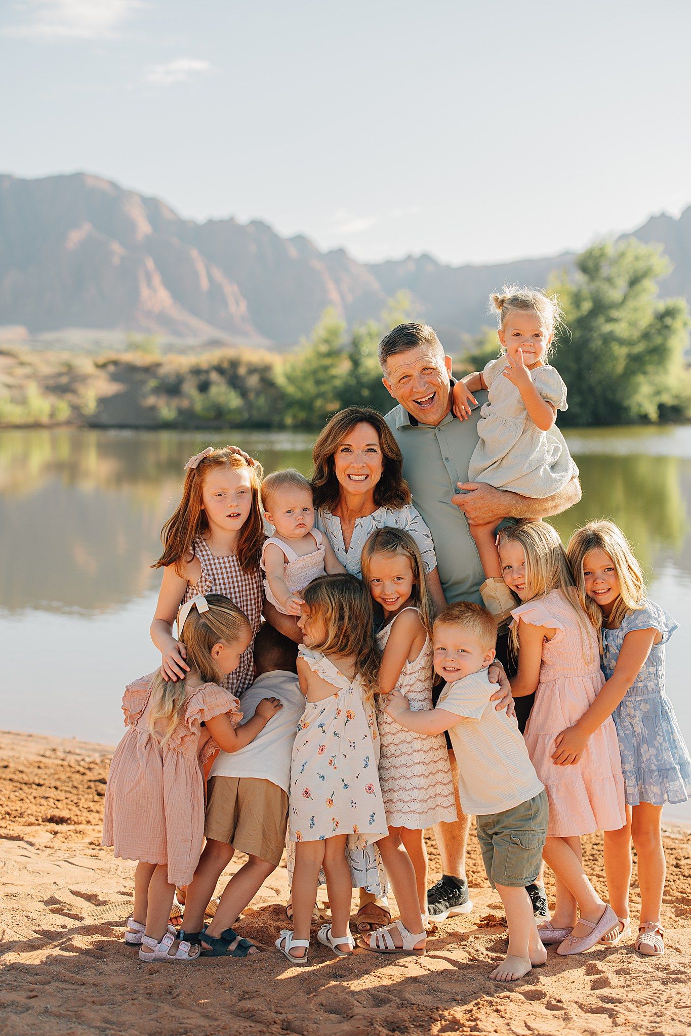 A large happy family with two parents and many children posing together on a sandy riverbank with mountains in the background.
