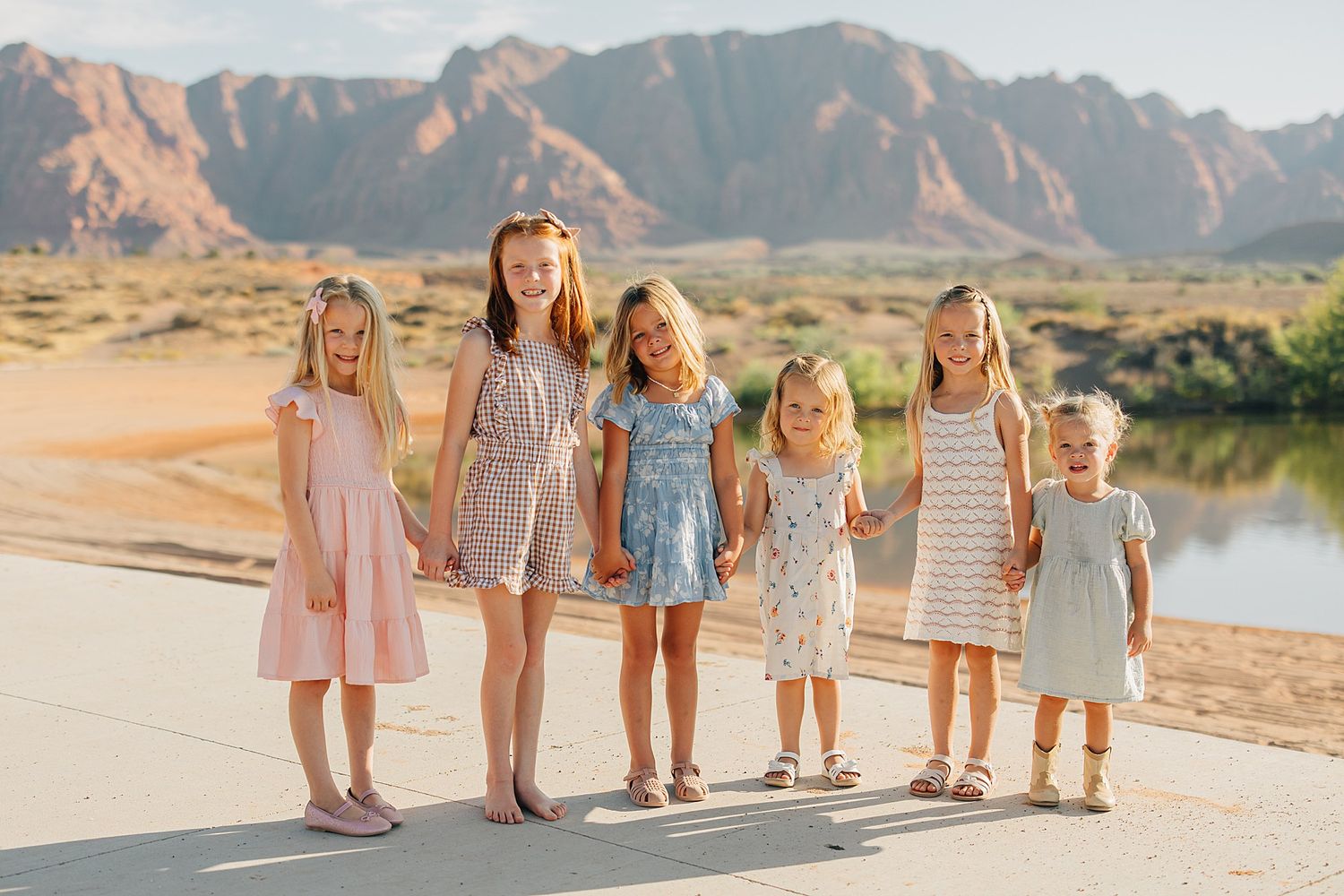 Six young girls in pastel dresses hold hands outdoors with scenic desert mountains and a river in the background.