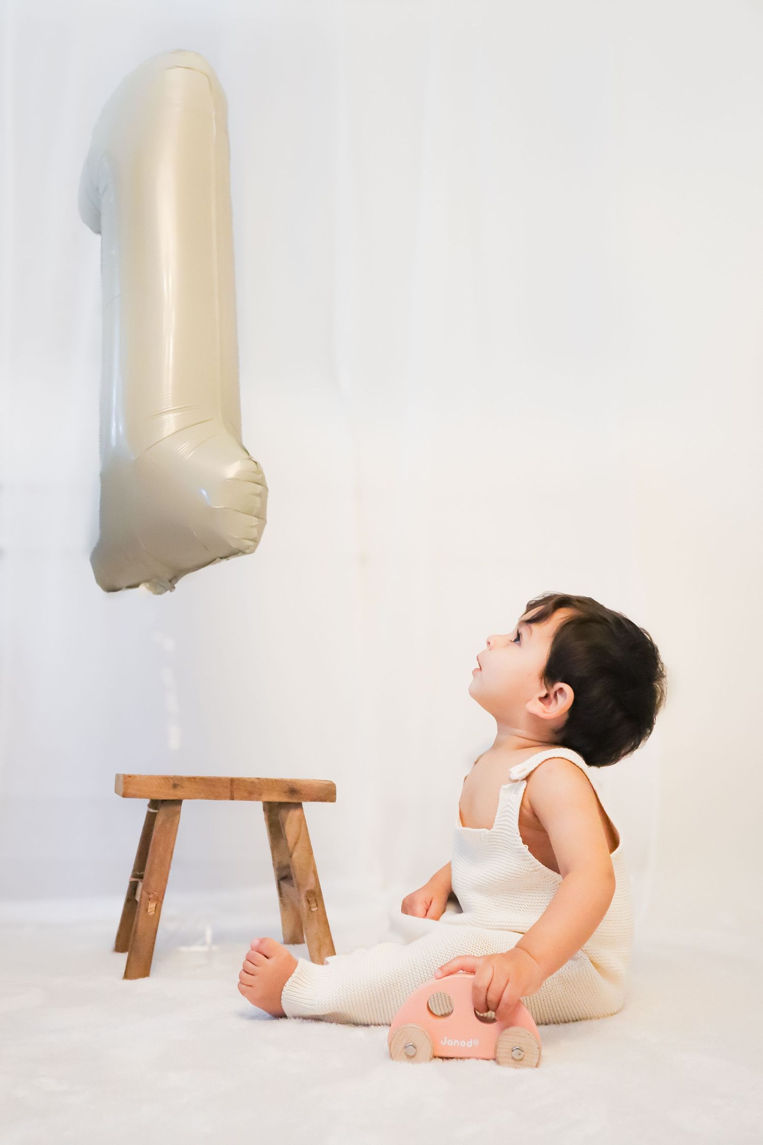 A baby in a white outfit sits on a fluffy rug, looking up at a number 1 balloon on their first birthday.