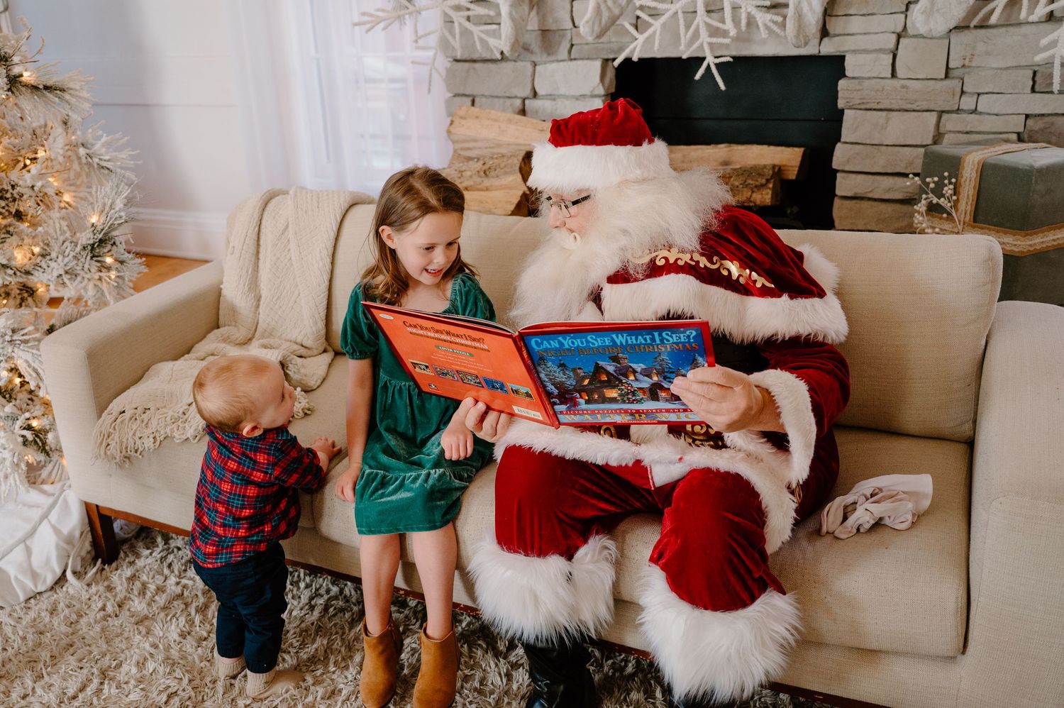 Santa Claus reads a colorful book to two young children on a cream sofa beside a decorated Christmas tree.