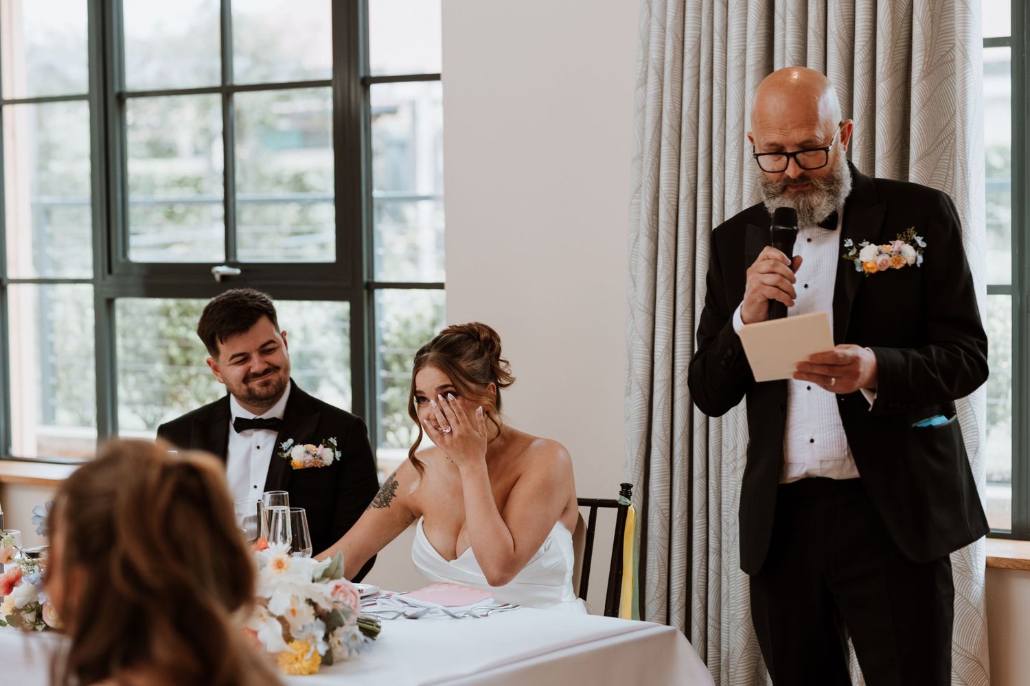 Father of the bride gives emotional wedding speech as bride covers her face and groom smiles at reception.