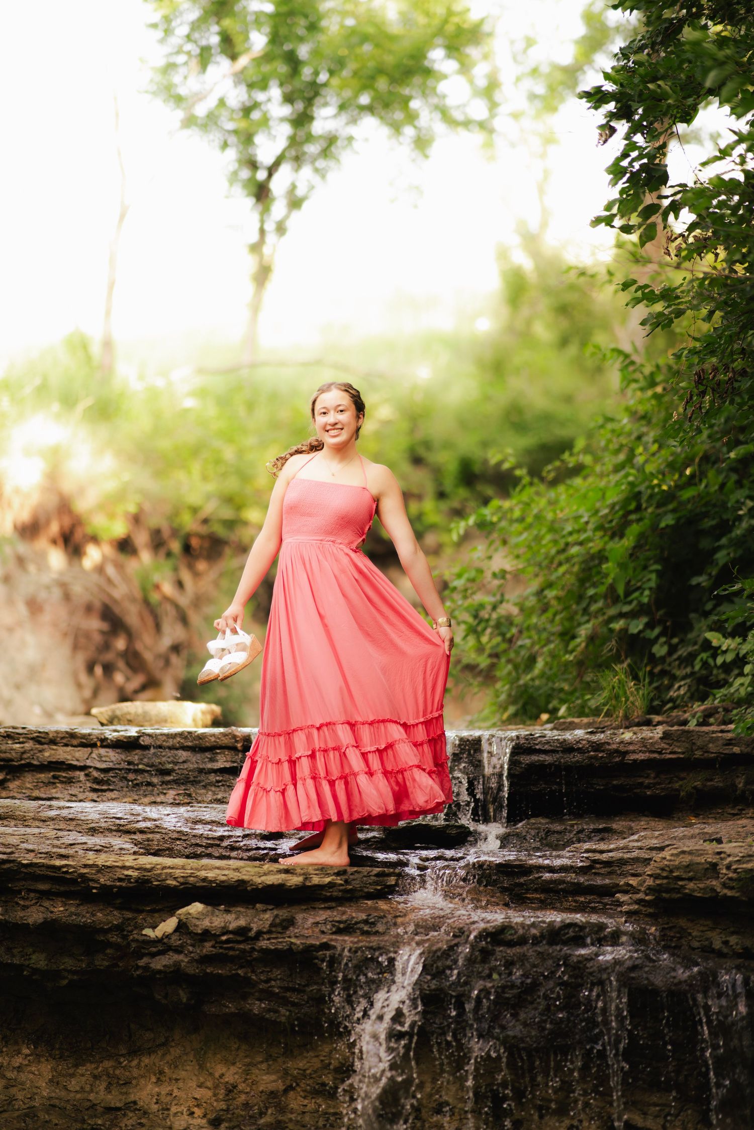 Young woman in coral maxi dress standing barefoot on rocky waterfall surrounded by lush green trees.