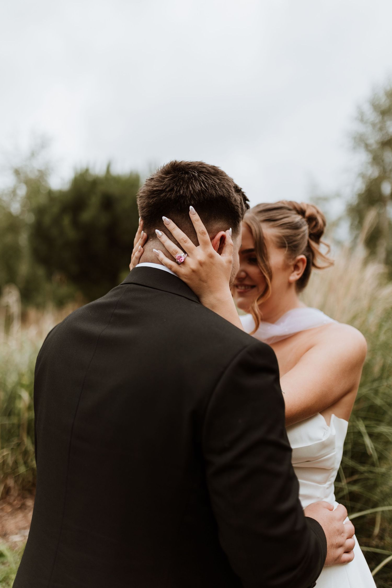 Bride in white halter gown smiles as she holds groom's face, showcasing her engagement ring outdoors.