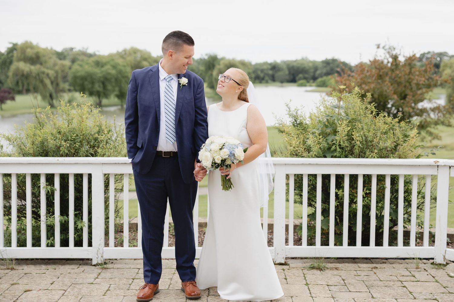 Bride holding white bouquet and groom in navy suit posing on terrace near pond.
