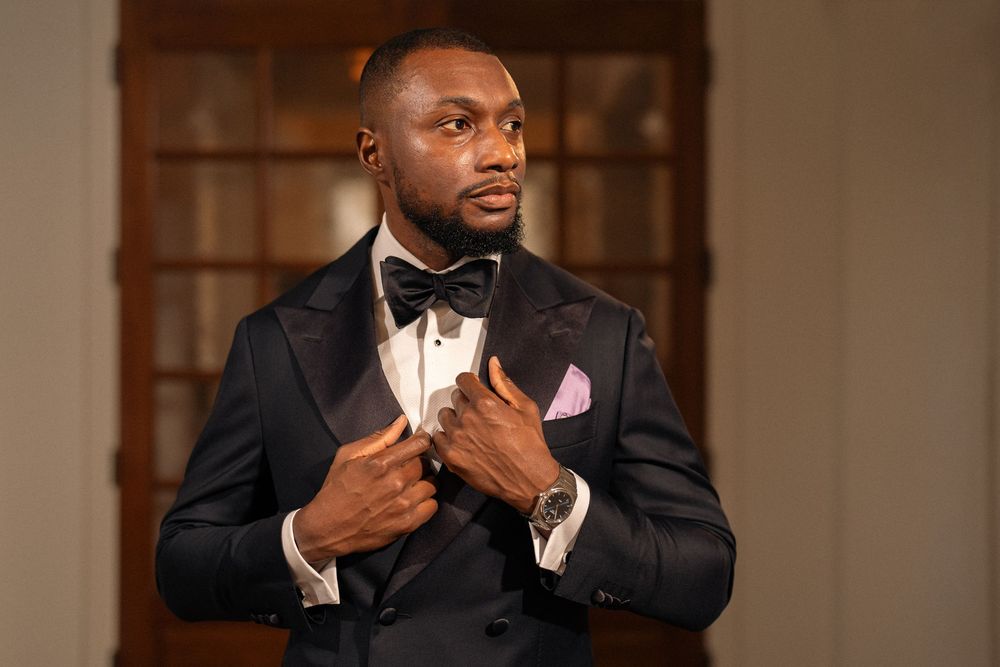 Elegant man in black tuxedo with bow tie and pink pocket square adjusting jacket in formal setting.