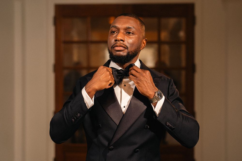 Elegant man in black tuxedo adjusting bow tie, formal attire with watch, indoor setting.