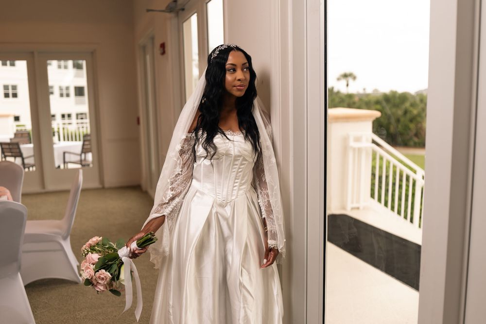 Elegant bride in white satin gown and veil holding pink bouquet standing in sunlit doorway.