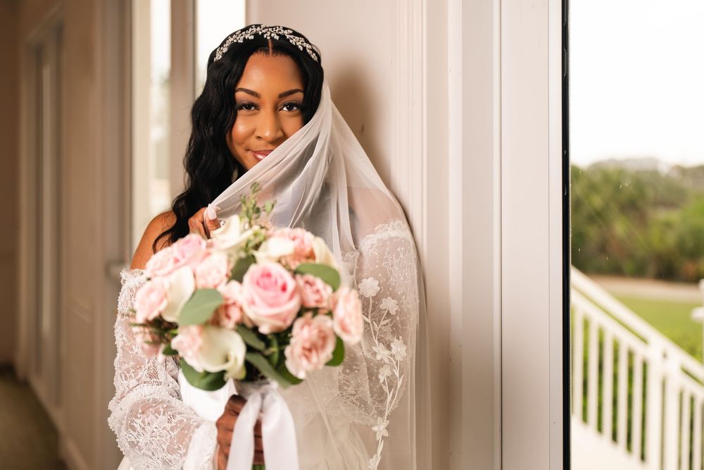 A beautiful Black bride in a lace wedding gown and veil holds a pink rose bouquet while posing by a window.