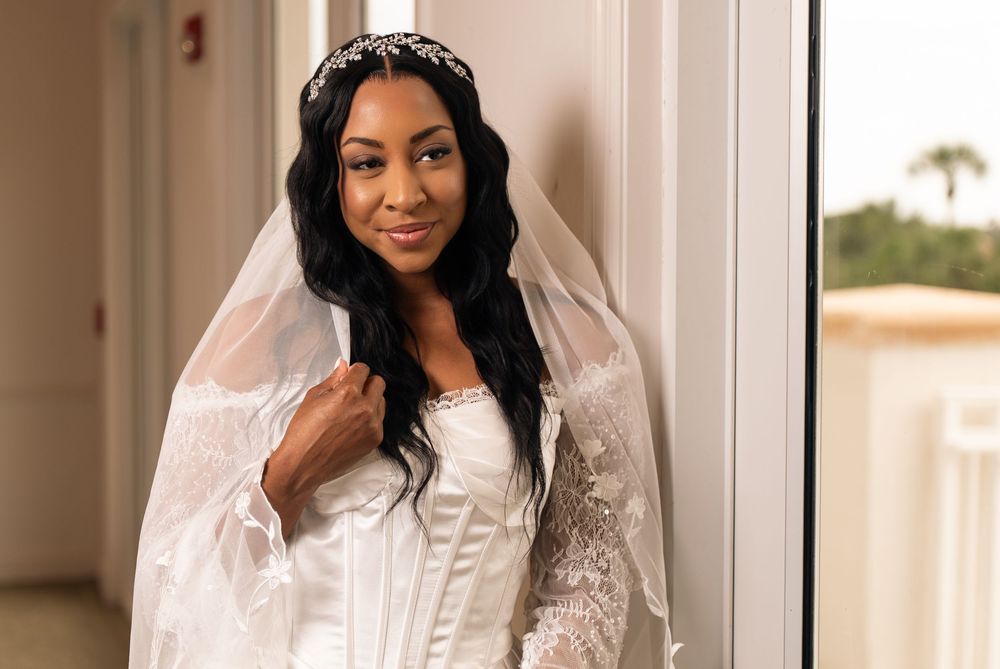 Elegant bride in white lace gown and veil wearing a crystal tiara, posing gracefully by a window.