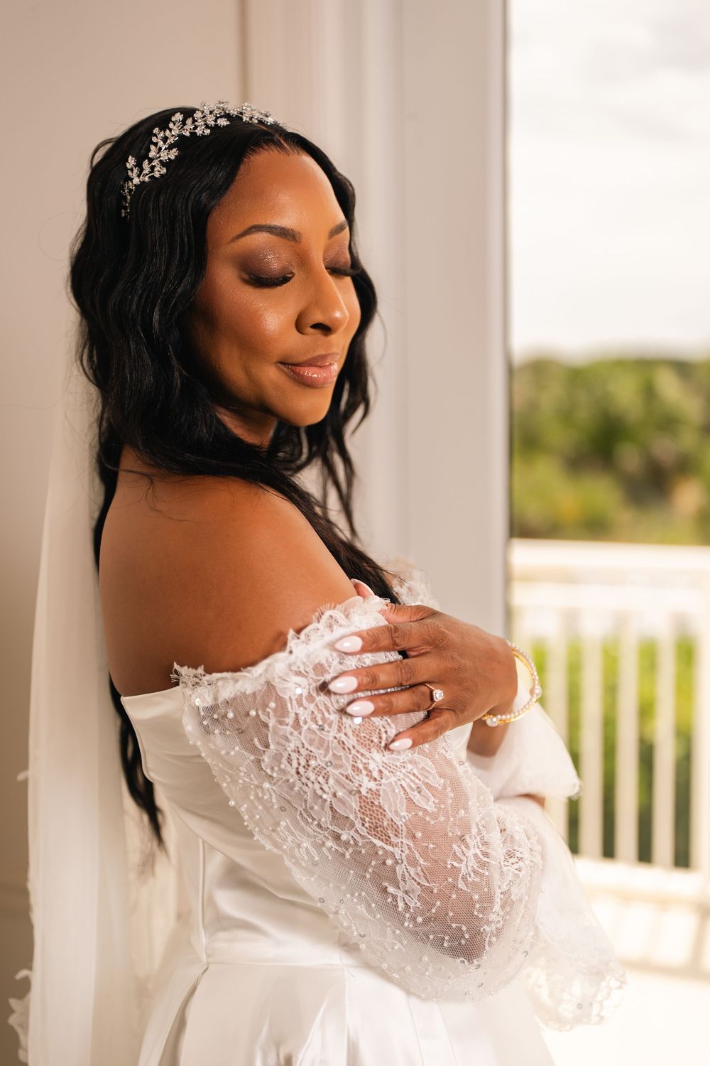 Elegant bride in off-shoulder lace wedding gown with pearl headpiece, eyes closed, posing near a sunlit window.