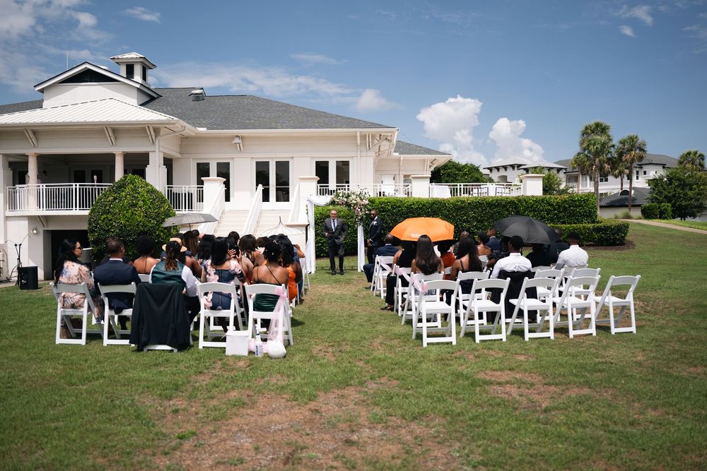 Outdoor wedding ceremony on green lawn beside white venue building with guests seated in white chairs under sunny skies.