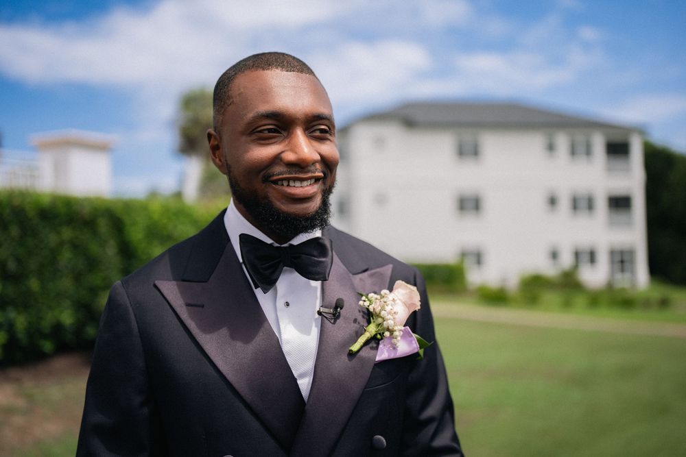 Smiling groom in black tuxedo with bow tie and floral boutonniere standing outdoors on wedding day.