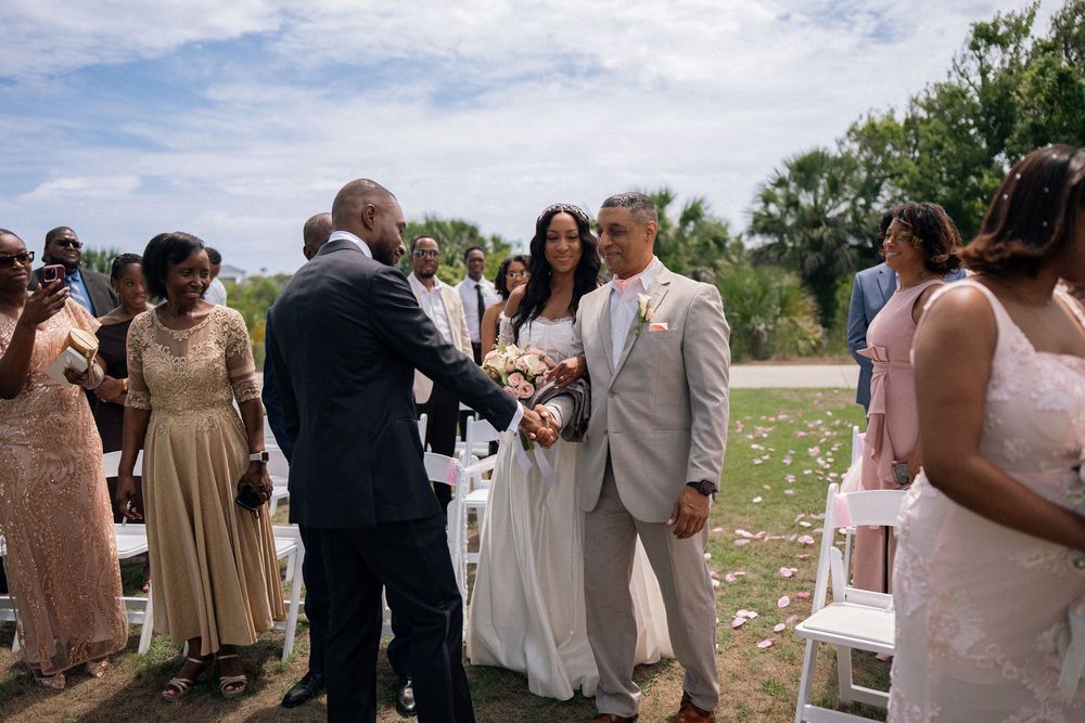 Outdoor wedding ceremony with bride and groom greeting guests along the aisle, surrounded by family in elegant attire.