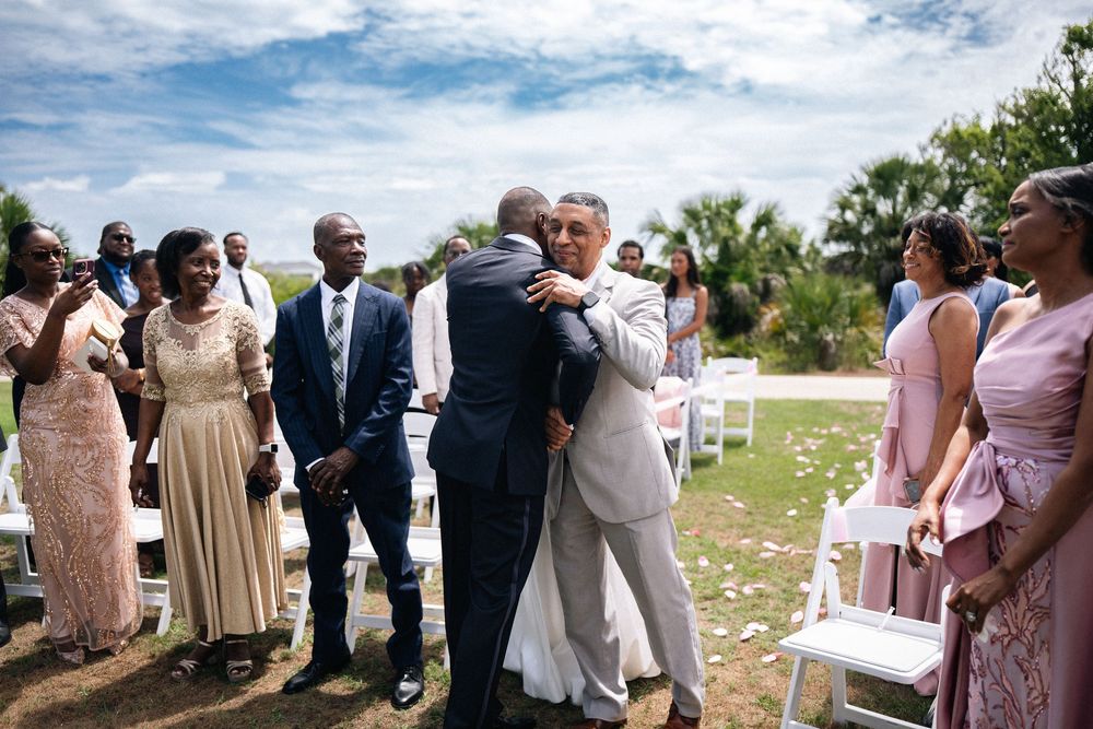 Two men share a heartfelt hug during an outdoor wedding ceremony while guests in formal attire look on.