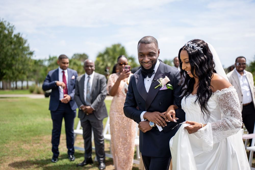 Smiling newlywed couple in wedding attire walking together outdoors with wedding guests in background.
