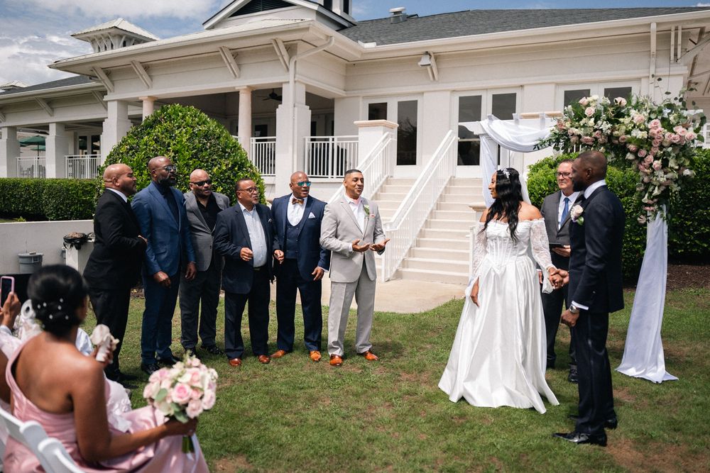 Outdoor wedding ceremony with bride and groom at floral arch, groomsmen in navy suits, elegant white venue backdrop.